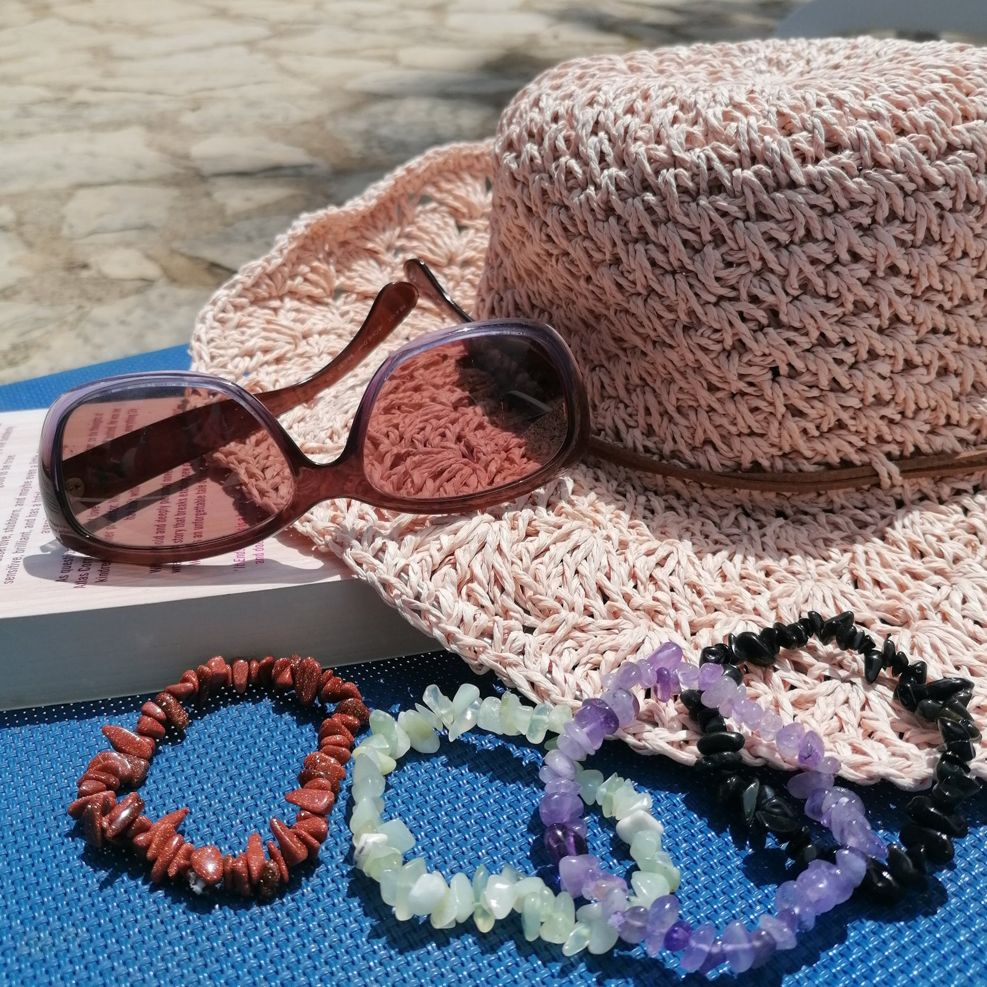 A straw sun hat with sunglasses and a book as the background and a goldstone, green new jade, purple amethyst and a black obsidian crystal chip bracelets