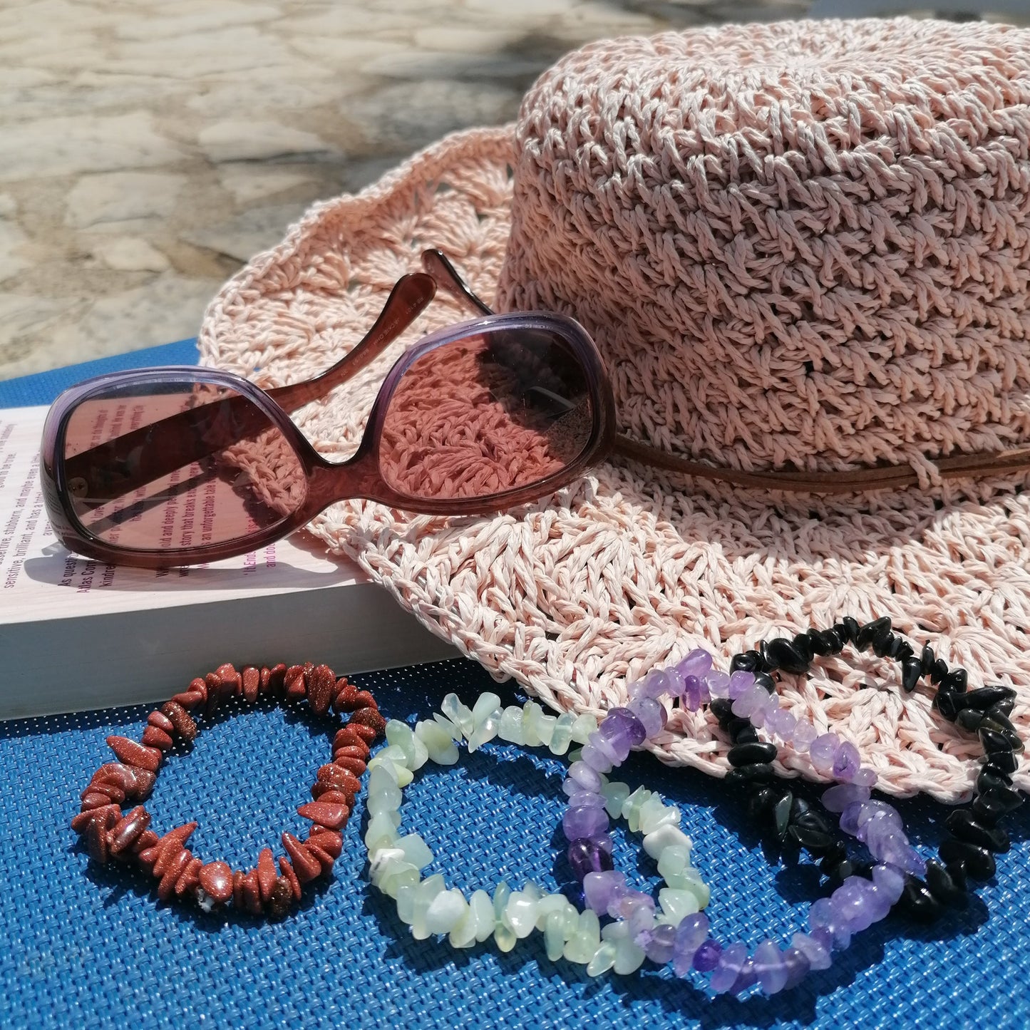 a straw sun hat, sunglass and a book background with a goldstone, a green new jade, an amethyst, and a black obsidian chip bracelets